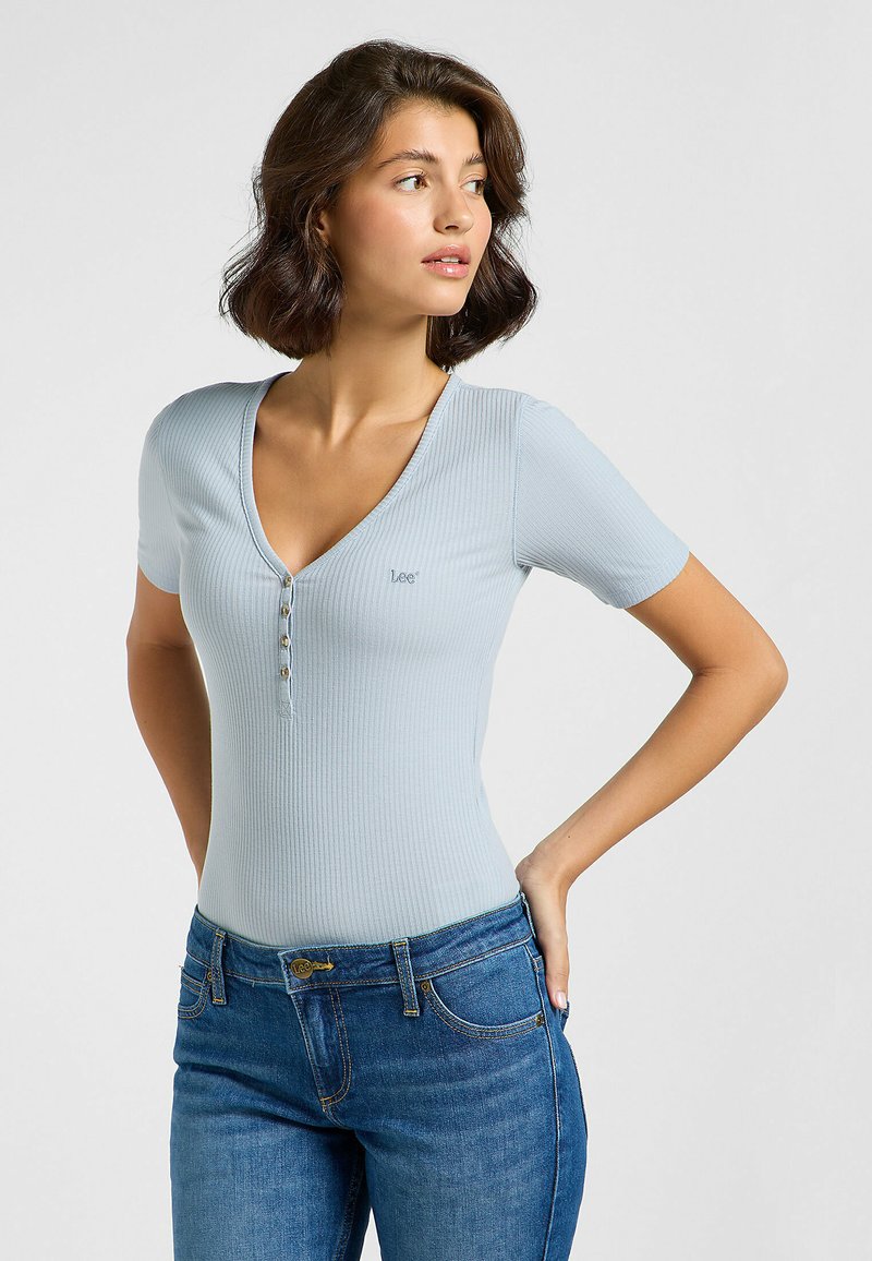 Body corto a maniche corte in blu chiaro a coste con scollo a V, caratterizzato da quattro bottoni frontali e un logo del marchio discreto. Indossato con jeans blu.