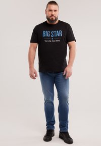 Habemeajaga mees, kellel on tagasikinnitatud juuksed, must "BIG STAR" t-särk, sinised teksad ja mustad tennised, seisab ühevärvilise tausta ees.