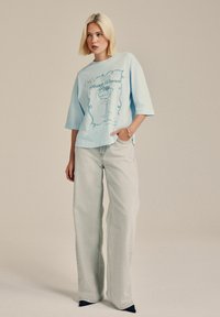 Blonde Frau in einem hellblauen, übergroßen Graphic-T-Shirt und weit geschnittenen, hellgrauen Jeans, steht mit einer Hand in der Tasche.