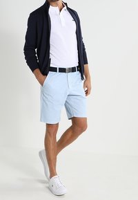 Cardigan bleu marine zippé sur un polo blanc, associé à un short bleu clair et des baskets blanches, avec une ceinture noire et une texture lisse.