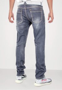 Graue Denim-Jeans mit Verblassungen, orangefarbener Stickerei und fünf Taschen, getragen mit schwarz-weißen Sneakers. Der Bund wird von einem gemusterten Gürtel ergänzt.