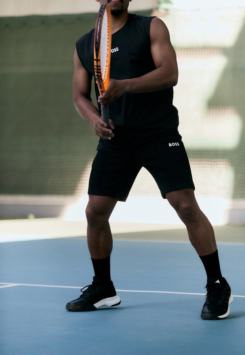 Cămașă fără mâneci de culoare neagră și pantaloni scurți cu logo-ul "BOSS", ținând o rachetă de tenis cu un cadru portocaliu, purtând încălțăminte sport neagră și șosete.