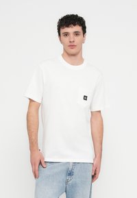 Λευκό βαμβακερό T-shirt με κοντά μανίκια, με τσέπη στο στήθος που φέρει μαύρη ετικέτα. Κανονική εφαρμογή με στρογγυλή λαιμόκοψη.