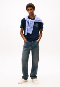 Marineblå T-skjorte i bomull med en logo på brystet, kombinert med en lys blå strikket genser båret casual over skuldrene, løstsittende denimjeans og hvite sneakers.