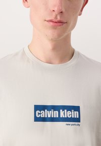 Lys grå bomull t-skjorte med en blå rektangulær logo med teksten "calvin klein" og "new york city" i hvitt, rund hals.