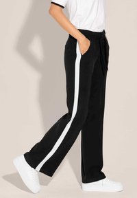 Zwarte wijde broek met een witte zijstreep, voorzien van een strik taille en zakken, gecombineerd met witte sneakers. Glad materiaal, comfortabele pasvorm.