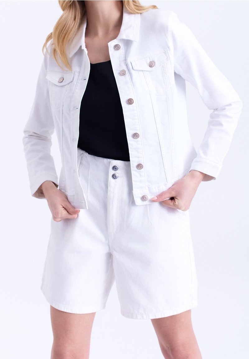 Bílá džínová bunda s měděnými knoflíky, dvěma náprsními kapsami a odpovídajícími bílými šortkami. Černá top vrstva viditelná pod tím. Hladká textura.