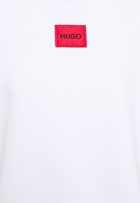 Sudadera blanca con un parche rectangular rojo que lleva la palabra "HUGO" en bordado negro. La tela es suave y ligeramente texturizada.