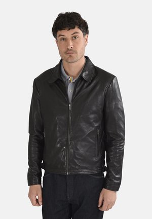 Homme portant une veste en cuir noire zippée par-dessus une chemise grise à col, debout devant un fond clair uni.