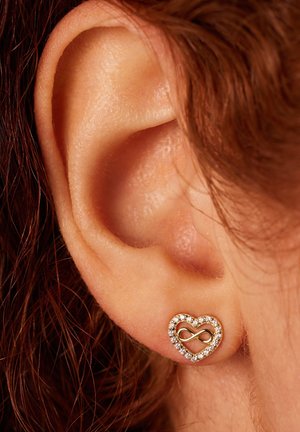 Boucle d'oreille en or en forme de cœur avec un symbole de l'infini au centre, entourée de petites pierres claires, présentée près de l'oreille.
