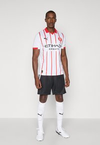 Maillot de football blanc avec des rayures verticales rouges, logo noir et col ; associé à un short noir, des chaussettes blanches avec le logo Puma et des crampons blancs.