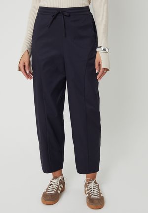 Femme portant un pantalon bleu marine fuselé avec taille à cordon, un pull côtelé beige, un bracelet manchette argenté et des baskets marron avec des lacets blancs.