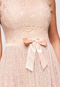 Robe sans manches en dentelle rose blush avec motif floral, taille cintrée, jupe en tulle à pois et nœud en satin noué à la taille.