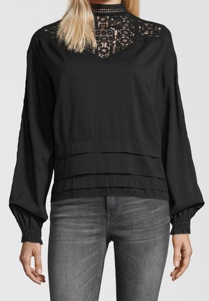 Mujer con blusa negra de manga larga con escote de encaje y dobladillo en capas, combinada con jeans ajustados grises, de pie contra un fondo liso.