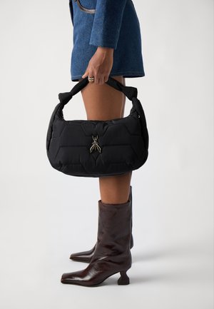 Borsa a tracolla nera trapuntata con motivo geometrico, dotata di un manico corto imbottito e di un charm a forma di insetto metallico; indossata con stivali marroni al ginocchio.