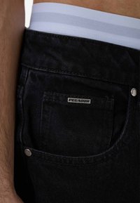 Granatowe dżinsy mają zbliżenie na tylnią kieszeń z srebrną etykietą z napisem "PEGADOR". Pasy mają białą i jasnoniebieską elastyczną taśmę w paski.