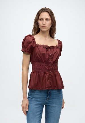 Frau mit welligem braunem Haar, die eine bordeauxrote Bluse mit Puffärmeln, Knöpfen und Spitze trägt, kombiniert mit blauen Jeans, schaut zur Seite.