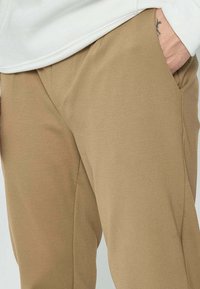 Personne portant un pantalon beige et un haut blanc, avec la main droite partiellement dans la poche et un petit tatouage visible sur le poignet.