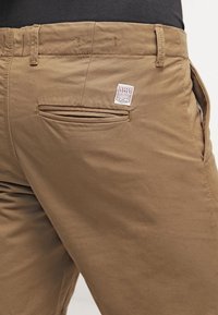 Pantalones cortos de lona marrón con un bolsillo trasero y una Etiqueta en la cinturilla. La tela parece suave con una textura agradable y costuras mínimas.