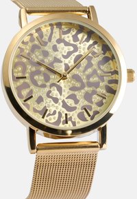 Ρολόι χρυσού τόνου με πρόσωπο σε λεοπάρ σχεδίαση που περιλαμβάνει γυαλιστερά αξεσουάρ. Ο αναδιπλούμενος ιμάντας σε χρυσό ολοκληρώνει το σχέδιο, αναδεικνύοντας το μοναδικό του μοτίβο.