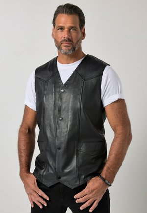 Mann mit Bart, der eine schwarze Lederweste über einem weißen T-Shirt trägt, steht mit den Händen nahe den Taschen vor einem einfarbigen Hintergrund.