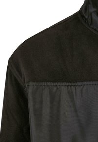 Svart jakke med en myk, fleece-lignende tekstur på den øverste delen og en glatt, blank overflate på den nedre delen, med kontrasterende sømmer.