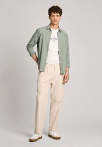 Camicia verde chiaro a maniche lunghe, abbottonata sopra una t-shirt bianca con grafica, abbinata a pantaloni crema e sneakers bianche con suola marrone. Vestibilità casual.