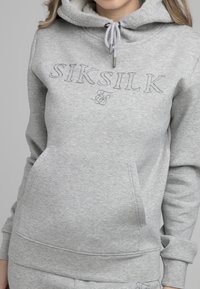 Mujer vistiendo una sudadera Siksilk gris claro con bolsillo frontal y capucha con cordón, mostrada de los hombros a las caderas sobre un fondo plano.