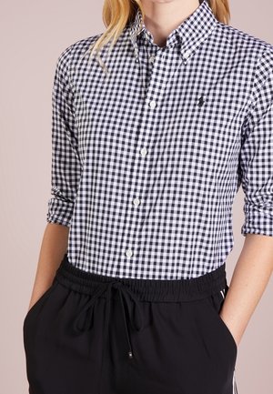 Camicia a quadri bianca e nera con colletto button-down, maniche arrotolate e un logo discreto sul petto. Abbinata a pantaloni neri.