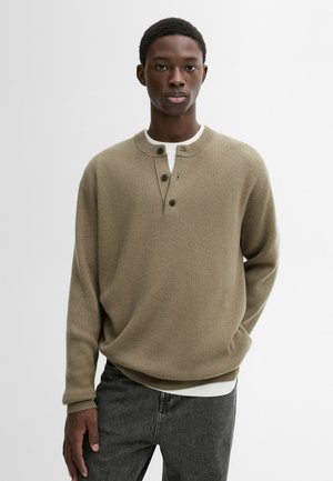 Pullover a coste color verde oliva con scollo henley a tre bottoni e maniche lunghe, indossato sopra una camicia bianca, abbinato a jeans scuri.