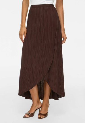 Une femme porte une jupe midi plissée taille haute marron foncé avec un ourlet asymétrique et des sandales à talons ouvertes marron.
