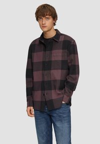 Chemise en flanelle à carreaux en violet foncé et noir, avec un col pointu, une poche avant et une fermeture à boutons. Associée à un jean bleu.