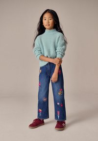 Suéter de punto texturizado en azul claro; jeans de pierna ancha en azul con bordado floral colorido; zapatillas color marrón con suela texturizada.