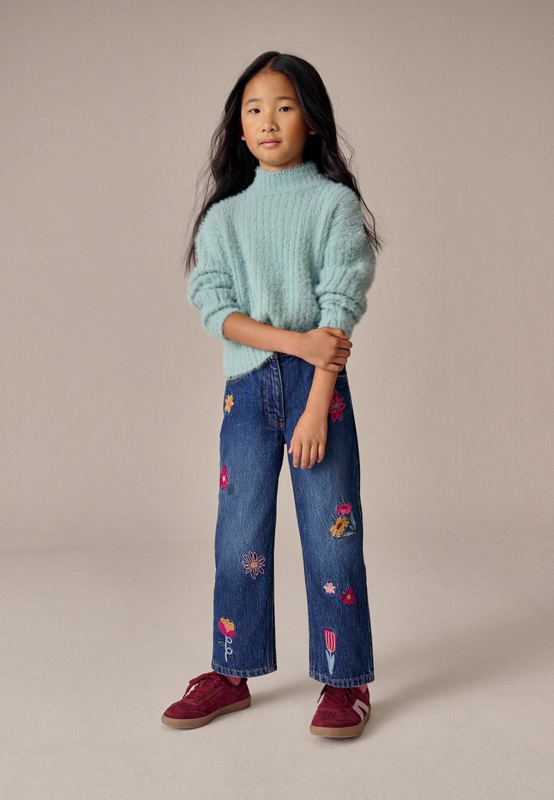 Maglione a maglia blu chiaro con texture; jeans larghi blu con ricami floreali colorati; sneakers bordeaux con suola texturizzata.