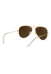 Occhiali da sole aviator dorati con lenti marroni, montatura in metallo sottile, para-naso in gomma e punte delle stanghette in plastica trasparente.