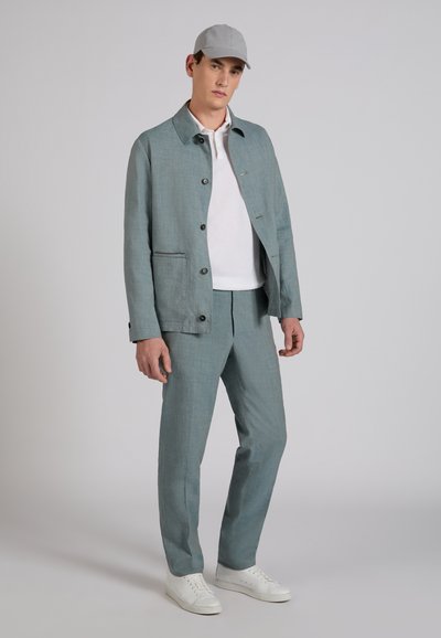 Homme en costume bleu clair avec veste et pantalon, polo blanc, casquette grise et baskets blanches, debout devant un fond uni.