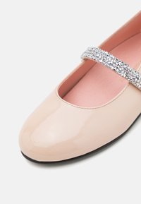 Zapato plano de charol beige brillante con puntera redonda y una correa adornada con strass plateados sobre el empeine. Suela negra, forro rosa.