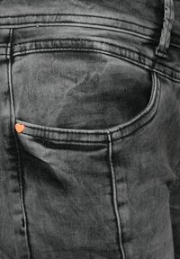 Svarta jeanstyg med en liten orange hjärta som sticker ut från bakfickan, med subtil blekning och synlig sömnad.