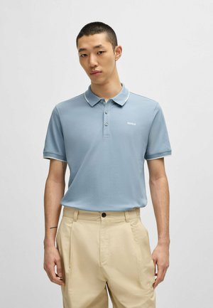 Light blue polo shirt with a standard collar, three-button placket, and small white logo. Paired with beige, pleated trousers.