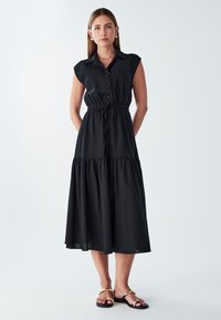 Czarna sukienka zapinana na guziki z kołnierzykiem, krótkimi rękawami, wiązaniem w talii i warstwowym dołem. Gładki materiał z luźnym krojem i długością sięgającą do podłogi.