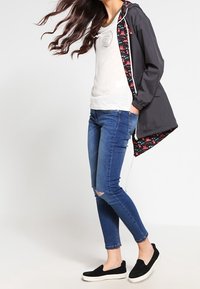 Czarna kurtka z kapturem z kwiecistym podbiciem, biała koszulka, postrzępione niebieskie dżinsy i czarne buty wsuwane. Włosy falowane; model stoi z boku.