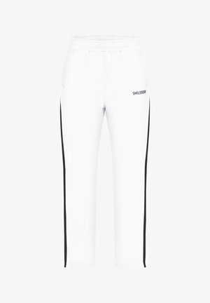 Homme en sweat à capuche blanc zippé et pantalon de survêtement blanc assorti avec des bandes latérales noires et des baskets blanches, debout avec les mains jointes.