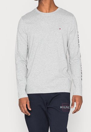 Homme portant une chemise manches longues gris clair avec un petit logo sur la poitrine et un pantalon bleu marine avec un logo sur la cuisse, debout devant un fond uni.