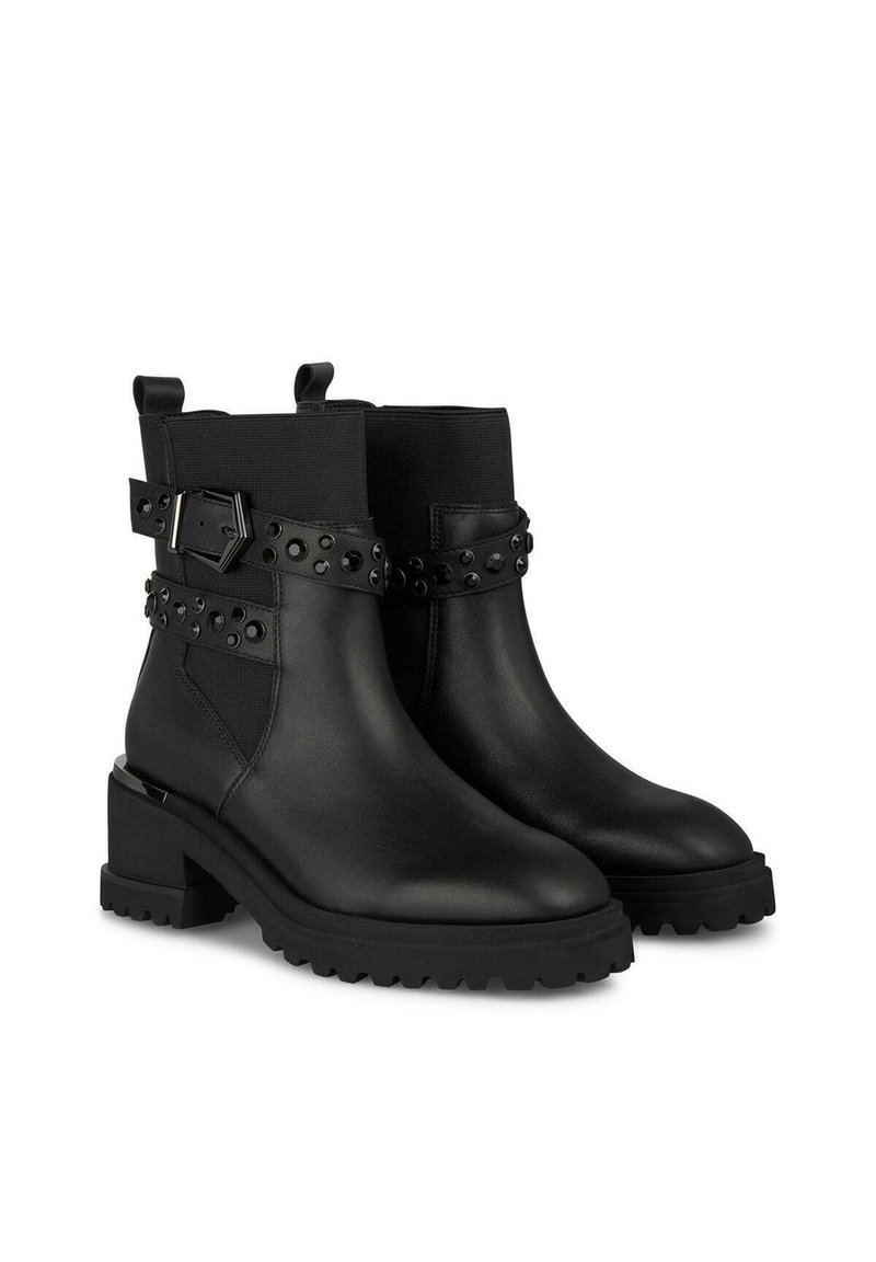 Botas de tobillo de cuero negro con suela de goma gruesa, que presentan una correa decorativa con tachuelas negras y un detalle de hebilla rectangular.