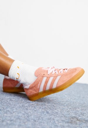 Osoba nosząca różowe buty Adidas Gazelle z białymi paskami i białe skarpetki na szarym wykładzinowym podłodze.