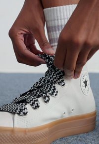 Witte canvas sneakers met een gum zool, zwart-witte houndstooth veters en een ronde logo patch aan de zijkant. Handen die de veters aantrekken.