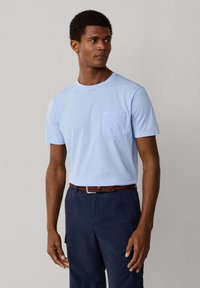 Homme portant un T-shirt bleu clair à manches courtes avec une poche frontale, rentré dans un pantalon bleu marine avec une ceinture marron, regardant sur le côté.
