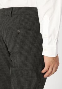 Pantaloni cu carouri gri închis, cu o textură netedă, având un singur buzunar pe spate cu închidere cu buton, stilizați cu o cămașă albă.