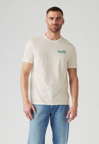 T-shirt crème à manches courtes en coton, avec un logo Levi's vert sur la poitrine gauche, associé à un jean en denim bleu clair.