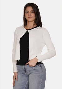 Cardigan bianco corto con maniche lunghe, scollatura rotonda e dettagli con bottoni, indossato sopra una maglietta nera, abbinato a jeans azzurri chiari.